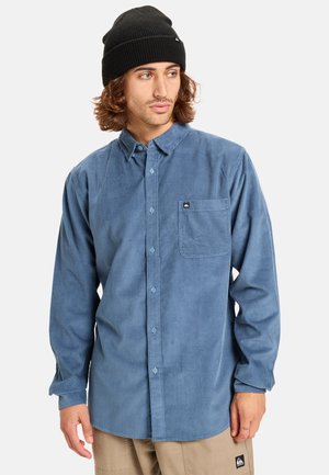 Camisa de pana azul con mangas largas, diseño de botones, bolsillo en el pecho y una etiqueta de logo. Combinada con un gorro de lana negro.