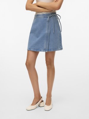 Kobieta nosząca niebieską, dżinsową spódnicę kopertową do kolan, zawiązaną z boku na kokardę, oraz kremowe sandały typu slingback na słupku. Ramiona skrzyżowane nad talią.