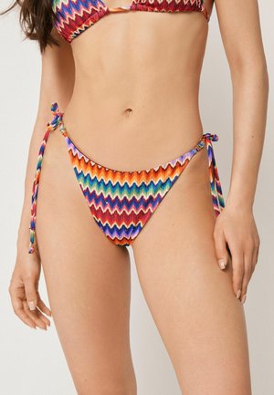 Kvinde iført en farverig bikiniunderdel med zigzag-mønster og sidestropper samt en matchende top, stående mod en ensfarvet baggrund.