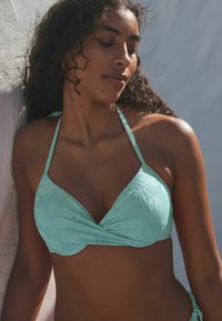 Menta zelena bikini gornji dio s trokutastim čašama, teksturiranim materijalom, uvijenim prednjim dijelom i tankim trakama, odložen na svijetloj pozadini.