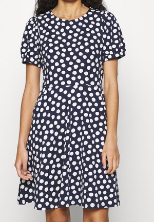 Femme portant une robe bleu marine à pois blancs irréguliers, avec des manches courtes bouffantes et une taille cintrée.