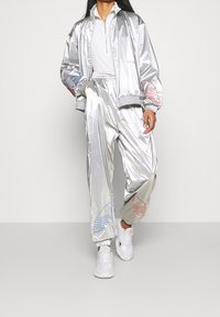Chándal plateado brillante con chaqueta con cremallera y pantalones con cintura elástica. Presenta diseños gráficos en rojo y azul en las mangas y las perneras.