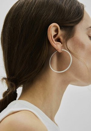 Žena s smeđom kosom vezanom unatrag, nosi veliki srebrni naušnicu u obliku obruča i bijelu majicu bez rukava, prikazana iz profila.