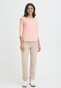 Pull rose pâle à manches trois-quarts avec des boutons noirs décoratifs sur l'épaule, associé à un pantalon chino beige et des baskets blanches.