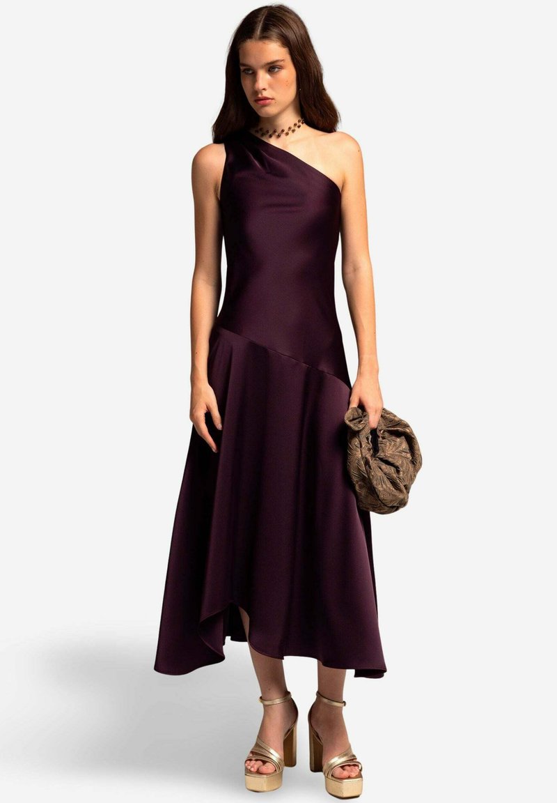Robe en satin violet asymétrique avec un corsage ajusté et une jupe fluide, associée à des talons compensés dorés et une pochette marron texturée.