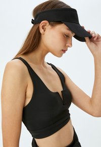 Top sportivo nero con scollatura a V bassa e spalline spesse, abbinato a un visore nero a tesa larga. Tessuto morbido, design aderente con lunghezza cropped.