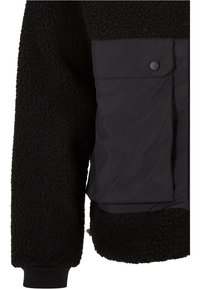 Giacca nera con un corpo in pile testurizzato e pannello in nylon liscio. Include una chiusura a bottoni a pressione e polsini a costine.