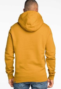 Žlutá mikina s kapucí, velkou kulatou kapucí, žebrovanými manžetami a spodním lemem. Tkanina se zdá být měkká a texturovaná. Žádné viditelné loga ani vzory.