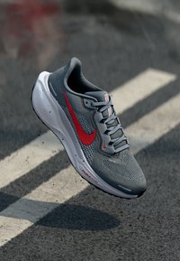 Gray athletic shoe with mesh upper, red Nike swoosh, white sole, and textured detailing. Features laces and branding on the side.