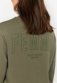 Alyvuogių žaluma džemperis su apvaliu iškirpimu. Nugarėlėje yra didelis užrašas "FEMME" ir žemesnėse eilutėse mažesnis logotipas "CREATIVE DEPARTMENT COPENHAGEN".