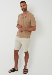 Camisa de manga corta de color marrón claro con textura y botones, combinada con pantalones cortos beiges y sandalias negras. Diseño sencillo con cuello y sin patrones.