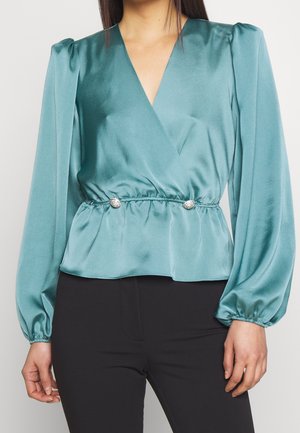 Vrouw draagt een teal satijnen wikkelblouse met lange opgepofte mouwen en twee decoratieve strassknopen bij de elastische taille, gecombineerd met een zwarte broek.