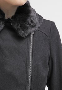 Svart ullkappa med en fuskpälskrage, asymmetrisk dragkedjestängning och mjuk textur, vilket framhäver en modern design.