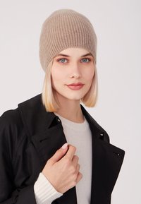 Beanie beige lavorato a maglia con texture a coste, indossato da una persona in un cappotto nero su un maglione leggero, che mette in mostra dettagli fatti a mano vicino al collo.