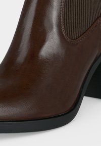 Bottine en cuir marron avec panneau latéral élastique et talon bloc noir, présentée en gros plan sur fond blanc.