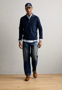 Polo a maniche lunghe blu navy sopra una camicia bianca con colletto, abbinato a jeans blu sbiaditi e sneakers marroni. Outift casual su uno sfondo neutro.