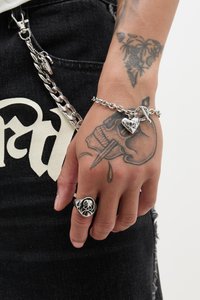 Sølvkjede armring med et hjertevedheng, båret på en hånd med en skalle-ring og fine tatoveringer. Svart denim bakgrunn med kontrasterende elementer.