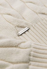 Tkanina w kolorze kremowym, dziana, z wzorami w warkocze oraz małą, srebrną metalkową etykietą z wygrawerowanym napisem "QUINTESSENCE".