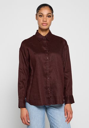 Žena s vlasy staženými dozadu, oblečená v tmavě hnědé košili s knoflíky a světle modrých džínách, stojí proti jednoduchému světle šedému pozadí.