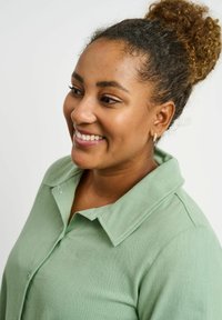 Green button-up shirt with a pointed collar, made of a textured fabric. Model has curly hair styled in a bun and wears small hoop earrings.