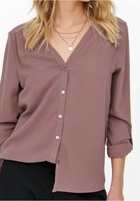 Camicia button-up di un leggero color malva con scollo a V, maniche a tre quarti e polsini arrotolati. Tessuto testurizzato con una vestibilità rilassata e bottoni in tono argento.