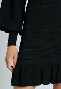 Abito nero lavorato a maglia con strisce testurizzate, dettagli a tessitura aperta e orlo a balze svasato. Maniche lunghe e voluminose con polsini a costine.