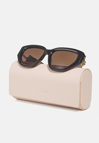 Black oversized sunglasses with brown gradient lenses placed on a textured light pink case, featuring a subtle logo embossing.