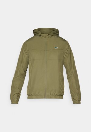 Olivgrön lättviktsjacka med dragkedja fram, huva och elastiska ärmmuddar. Har en horisontell söm och en liten broderad logotyp på bröstet.