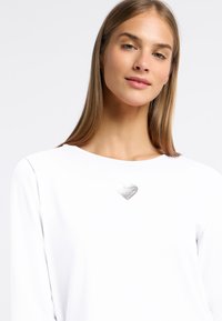 Weißes Langarmhemd mit einem silbernen Herz-Akzent auf der Brust. Glatte Textur und runder Ausschnitt, mit sichtbaren Nahtdetails.