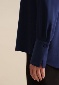 Camicia blu navy con una texture liscia, maniche lunghe e polsini con bottone. L'orlo è leggermente più lungo sul retro.