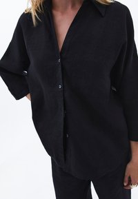 Czarna lniana koszula o luźnym kroju, z długimi rękawami i dekoltem w kształcie litery V. Posiada jedną kieszeń na piersi i zapięcie na guziki. Miękka tekstura.
