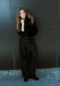 Czarna i zielona kurtka z futerkiem imitującym futro z szerokim kołnierzem, zestawiona z czarnymi, szerokimi jeansami z wysokim stanem. Modelka opiera się o ścianę.