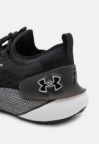 Baskets Under Armour en noir et blanc avec tissu en maille, semelle rembourrée, languette de traction au talon et détail du support de talon marqué.