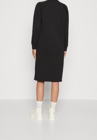Schwarzes knielanges Kleid mit langen Ärmeln, Raglan-Schultern, seitlichen Schlitzen und gerippter Textur. Kombiniert mit weißen Sneakern und Socken.