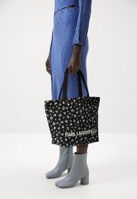 Sac fourre-tout noir avec logo blanc et petits motifs imprimés. Dispose de deux poignées noires robustes et d'un design rectangulaire spacieux.