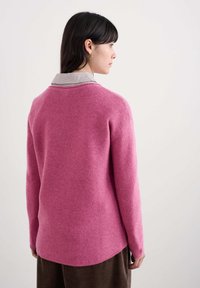 Różowy sweter dzianinowy o luźnym kroju, okrągłym dekolcie i długich rękawach. Teksturowany materiał z nieco dłuższym tyłem, nałożony na koszulę w paski.