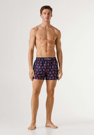 Młody mężczyzna stojący boso, ubrany w granatowe spodenki kąpielowe z czerwonym nadrukiem homara na jednolitym białym tle.