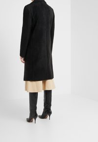 Abrigo de lana negro y texturizado con corte recto, mangas largas y cuello, combinado con botas negras hasta la rodilla y una falda beige.
