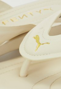 Light cream flip-flops with a smooth texture, featuring a gold Puma logo and stitching details on the straps.