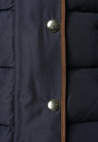 Marineblaue Steppweste mit glatter Oberfläche, braunen Akzenten und glänzenden silbernen Druckknöpfen an der Vorderseite zur Verschluss.