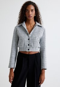 Harmaa tweed-tyylinen lyhyt takki, jossa on kaulus, nappikiinnitys ja pitkät hihat. Takissa on teksturoitu kuvio ja vyötärö on naisellinen.