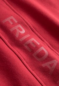 Rode stof met een zachte textuur heeft het woord "FRIEDA" gedrukt in een lichtere tint. Het ontwerp heeft opvallende stiksels langs de naden.