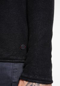 Schwarzer, strukturierten Pulloverärmel mit geripptem Bündchen; verfügt über ein kleines, rundes Logopatch mit rotem Akzent nahe der unteren Naht.