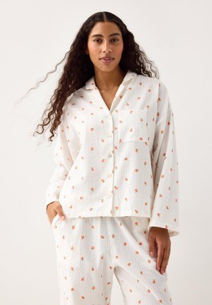Vrouw draagt een wit, gestructureerd pyjamapak met een klein oranje fruitpatroon, staand tegen een effen lichte achtergrond.