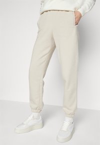 Beige joggebukser laget av myk stoff har elastiske mansjetter og sidelommer, parret med hvite sneakers. Enkelt og funksjonelt design.