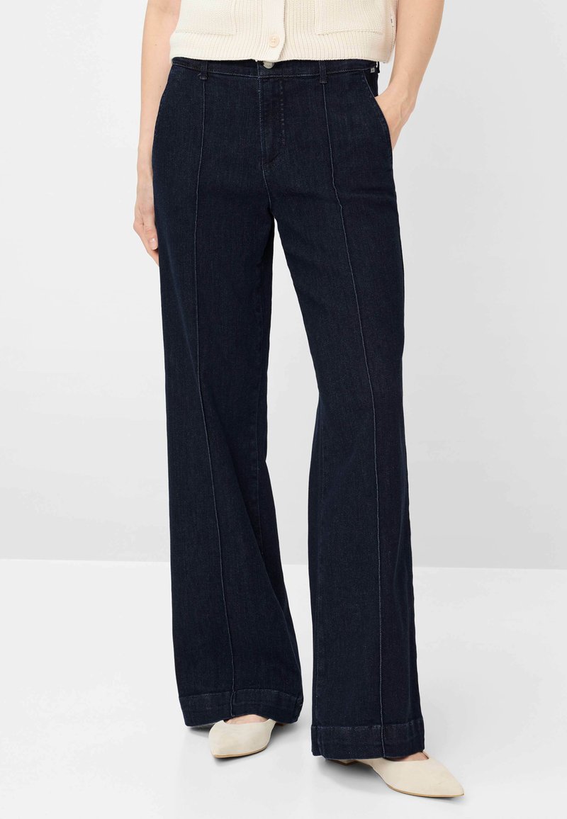 Donna con jeans larghi a gamba larga blu scuro, top senza maniche beige chiaro e ballerine beige chiaro con punta, in piedi con una mano in tasca.