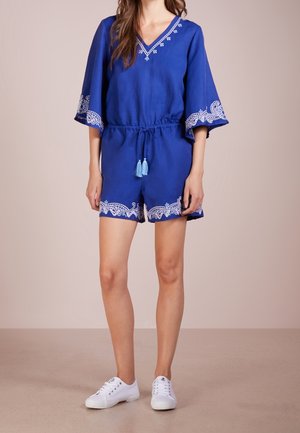 Femme portant une combinaison courte bleu à col en V avec des motifs brodés blancs et des pampilles, associée à des baskets blanches à lacets sur un fond neutre.