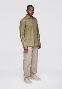 Chemise boutonnée vert olive clair avec une poche poitrine, associée à un pantalon beige et des chaussures de couleur claire. Fond uni.