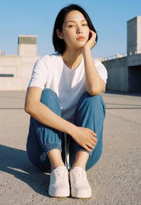 Fiatal nő rövid fekete hajjal, keresztbe tett lábbal ül betonon, fehér pólót, kék nadrágot és fehér tornacipőt visel, szabadban, napfényben.