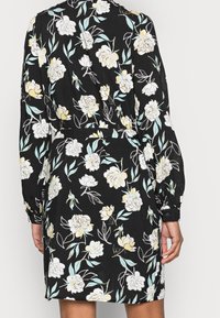 Černé květinové šaty s dlouhým rukávem, na kterých jsou bílé a světle žluté květy a světlomodré listy, které nosí stojící osoba viděná zezadu.